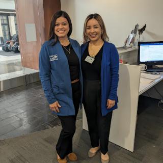 Two professionals posing together in an office setting, both wearing blue jackets.