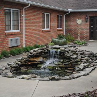 A serene stone-bordered pond with a cascading waterfall located in a courtyard next to a brick building.