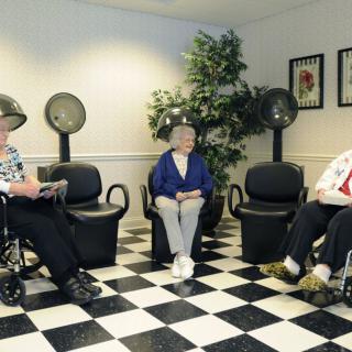 Three elderly women sit under hair dryers in a salon with checkered flooring.