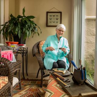 An elderly woman engaged in a crafting activity in a cozy indoor space, surrounded by sunlight and plants.