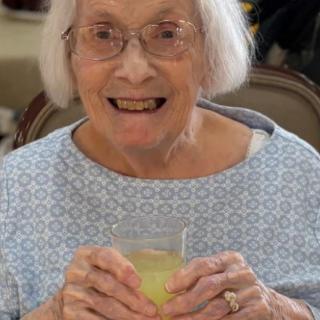 An elderly woman with glasses smiling and holding a drink indoors.