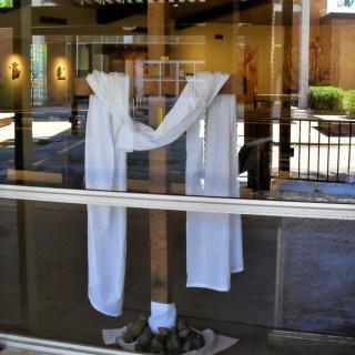 A wooden cross draped with a white cloth, seen through a glass window reflecting the interior.