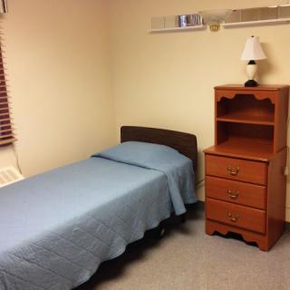 A simple, minimalist bedroom with a single bed, dresser, and lamp.
