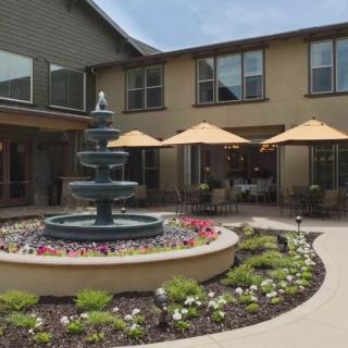 A courtyard featuring a central fountain surrounded by flowers, with outdoor seating shaded by umbrellas in front of a two-story building.