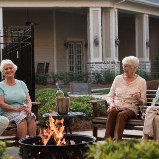 A group of seniors enjoying a gathering around a firepit in an outdoor garden setting.