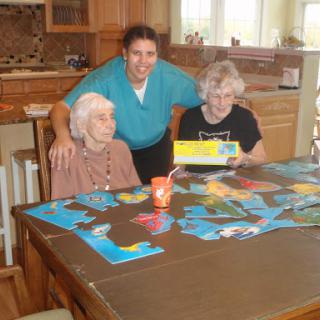 A caregiver enjoys a leisurely moment with two elderly women while playing a board game in a cozy kitchen setting.