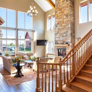 A spacious living room featuring a high ceiling with a wooden staircase, stone fireplace, and large windows letting in ample natural light.