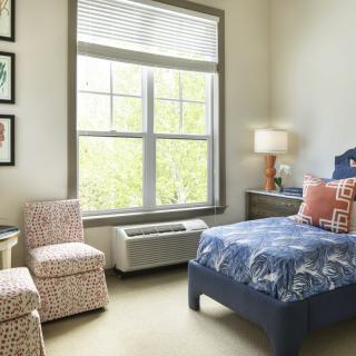 A cozy bedroom with a large window, featuring a neatly made bed and comfortable chairs.