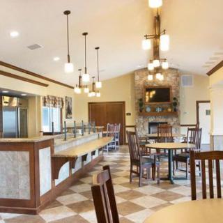 A cozy restaurant dining area featuring wooden tables and chairs, elegant lighting fixtures, a checkered floor, and a central stone fireplace.