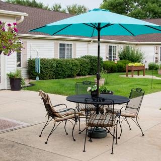 A cozy backyard patio with a table and chairs set under a teal umbrella, surrounded by lush greenery.