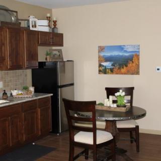 A cozy kitchen with wooden cabinets and a small dining table set for two, featuring a fridge and wall artwork.