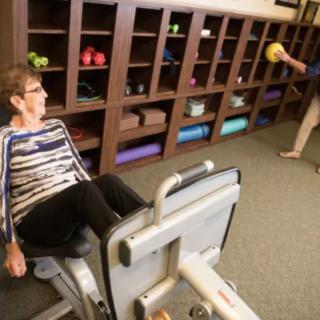An elderly woman uses a leg press machine in a gym setting, with exercise equipment neatly organized on shelves in the background.