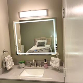 A modern bathroom vanity with a lighted mirror, reflecting a neatly made bed in an adjoining room.