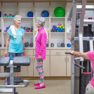 Two senior women chatting in a gym, surrounded by exercise equipment and colorful workout gear.