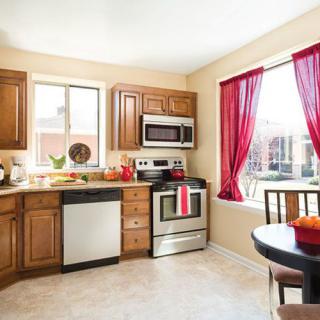 A brightly lit kitchen with wooden cabinets and stainless steel appliances, featuring a small dining area by the window with red curtains.