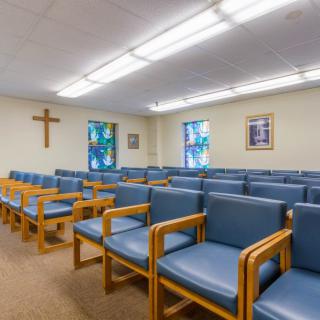 An interior view of a chapel with rows of blue cushioned chairs and a wooden cross on the wall.