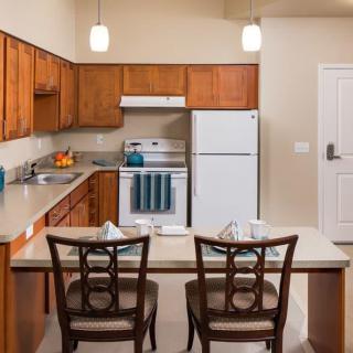 A modern kitchen with wooden cabinets, featuring a stove, refrigerator, and a small dining area with two chairs.