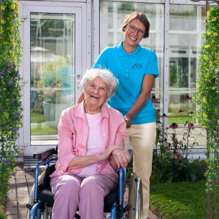 An elderly woman in a wheelchair is accompanied by her caregiver in a garden setting.