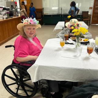 An elderly woman in a pink shirt and floral hat enjoys a meal in a dining area.