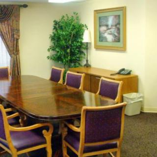 A small meeting room with a wooden conference table and upholstered chairs, featuring an indoor plant, artwork on the wall, and a telephone.