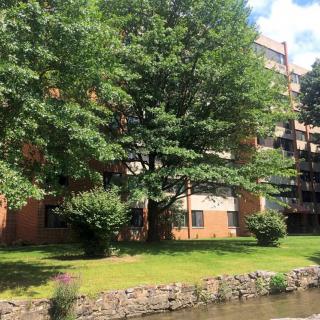A view of an apartment building surrounded by lush greenery and a nearby river on a sunny day.