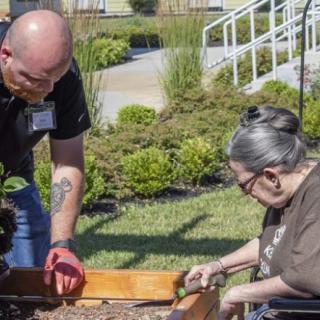 A volunteer assists an elderly person with gardening in a community garden.