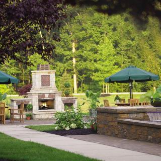 A serene garden patio with a stone fireplace, surrounded by lush greenery and shaded seating areas.
