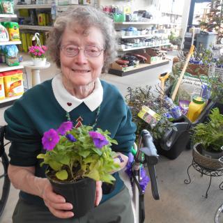 An elderly woman smiling while holding a potted plant with purple flowers at a garden center.