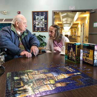 Two elderly individuals engage in conversation while working on a puzzle together.