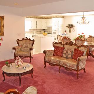 A cozy living room with vintage-style furniture and an open kitchen, featuring a red carpet and elegant chandelier.