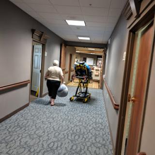 An elderly person walks down a hospital corridor while a healthcare worker assists a patient in a wheelchair.