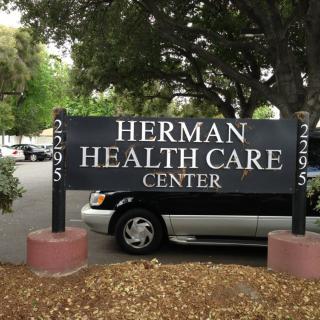 Sign for Herman Health Care Center in a parking area surrounded by trees.