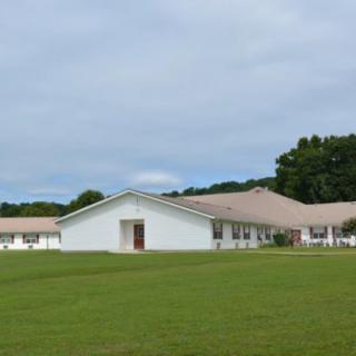 A single-story building surrounded by a grassy field and trees under a cloudy sky.