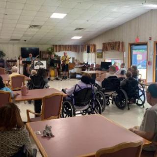 A community room in a nursing home with elderly residents gathered together, some seated in wheelchairs.