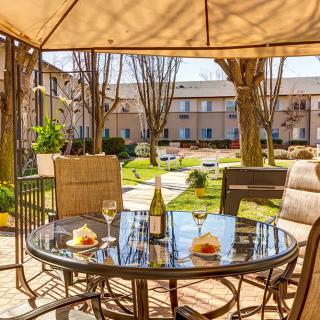 A cozy outdoor patio setup with a table and chairs under an umbrella, featuring wine glasses and a bottle, overlooking a well-maintained garden in an apartment complex.