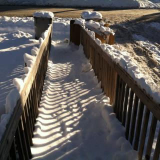 A snow-covered pathway lined with wooden fences casting shadows in the winter sunlight.