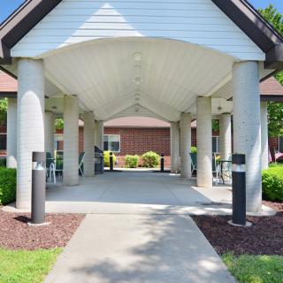 A covered walkway with columns leading to a building entrance, surrounded by landscaped greenery.