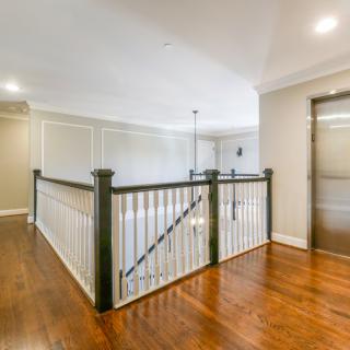 A well-lit hallway with hardwood flooring, featuring a staircase with a dark banister and an elevator.