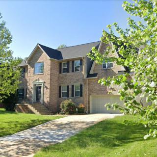 A large brick suburban house with a driveway and lush greenery on a sunny day.