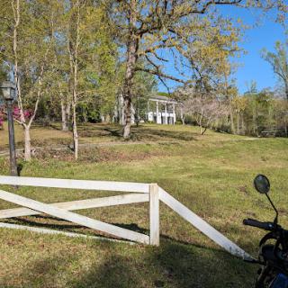 A scenic view of a rural landscape featuring a white gate, a motorcycle, and a house nestled among trees under a clear blue sky.