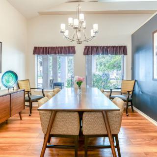 A bright and elegant dining room featuring a long wooden table, chairs, and a chandelier, with large windows letting in natural light.
