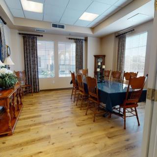 A well-lit dining room with a wooden floor, featuring a large table surrounded by chairs and adorned with curtains and windows.