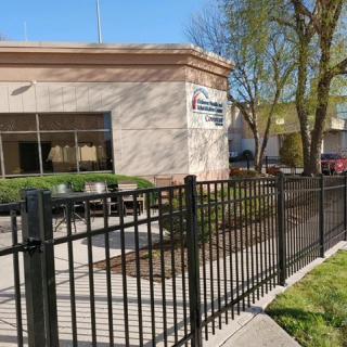 A small building with a black metal fence in front, surrounded by trees on a sunny day.