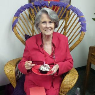 An elderly woman in a red outfit is smiling while holding a cupcake, seated indoors on a decorative chair.