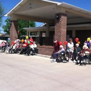 A group of elderly people in wheelchairs gathered outside a nursing home on a sunny day, holding colorful balloons.