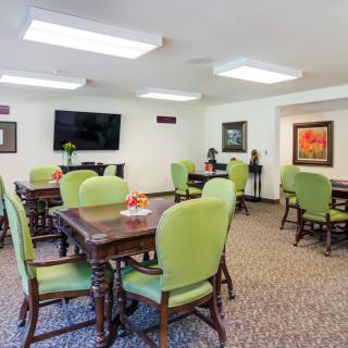 A well-lit dining area featuring green upholstered chairs, wooden tables, and a television on the wall.