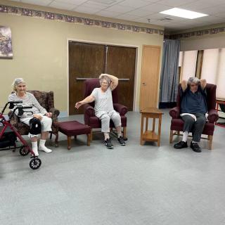 Three seniors exercising indoors with a walker and chairs.