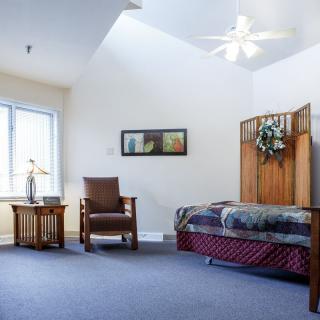 A cozy bedroom with a single bed, chair, and window under high ceilings and a ceiling fan.