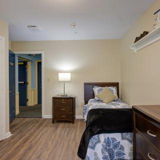 A cozy bedroom with a single bed and wooden furniture, featuring a lamp and simple decor.