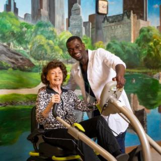 An elderly woman participates in a physiotherapy session with a healthcare professional assisting her on exercise equipment.